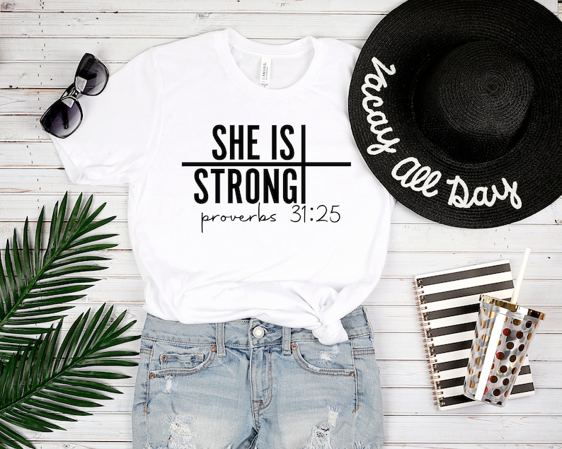 May include: White t-shirt with black text that reads "She is Strong" with a line through the word "Strong" and the text "Proverbs 31:25" below. The t-shirt is worn with denim shorts and a black sun hat with white text that reads "Vacay All Day".
