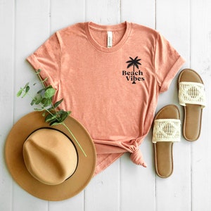May include: A coral pink t-shirt with a palm tree and the text "Beach Vibes" printed on the front. The t-shirt is laid out on a white wooden surface with a brown hat and two white sandals.
