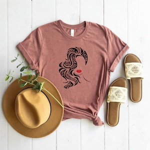 May include: A mauve t-shirt with a black silhouette of a woman's face with the words "fuerte", "poderosa", "mujer", "trabajadora", "alma", "latina" surrounding the face.
