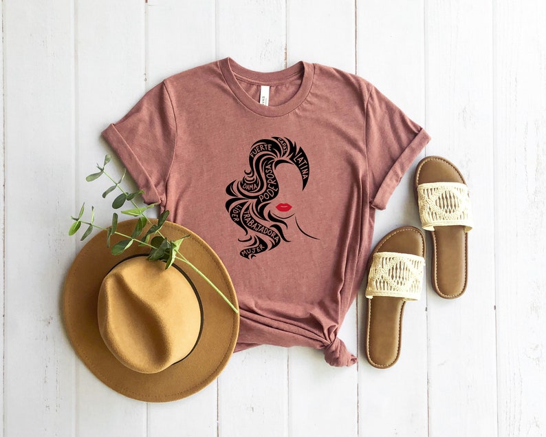 May include: A mauve t-shirt with a black silhouette of a woman's face with the words "fuerte", "poderosa", "mujer", "trabajadora", "alma", "latina" surrounding the face.