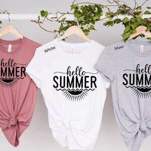 May include: Three t-shirts with the text "hello summer" in black lettering on a white background. The t-shirts are displayed on hangers and are in the colors mauve, white, and silver.