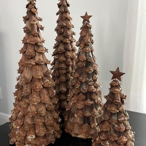 May include: Four decorative Christmas trees made from pinecones. The trees are various sizes and have a rustic brown color. Each tree is topped with a rusty metal star. The trees are arranged on a black surface.