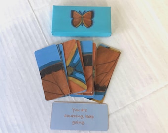 May include: A set of affirmation cards with a blue background and butterfly illustrations. The cards feature the text "You are amazing, keep going."