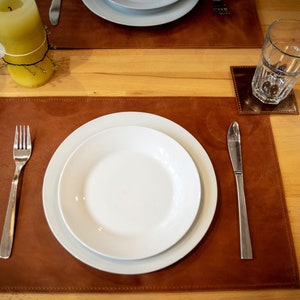 May include: A brown leather placemat with a white dinner plate, a fork, and a knife. The placemat is on a wooden table.