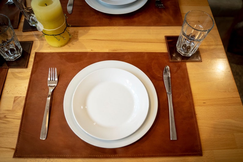 May include: A brown leather placemat with a white dinner plate, a fork, and a knife. The placemat is on a wooden table.