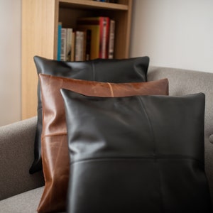 Pu&ograve; includere: Tre cuscini decorativi, un cuscino in pelle marrone con un cuscino in pelle nera sopra, e un cuscino in pelle nera sopra quello. I cuscini sono su un divano grigio.
