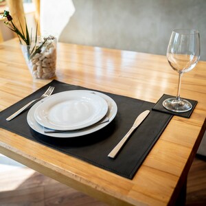 May include: A black leather placemat with a white plate, fork, and knife. A glass of water sits on a matching leather coaster.