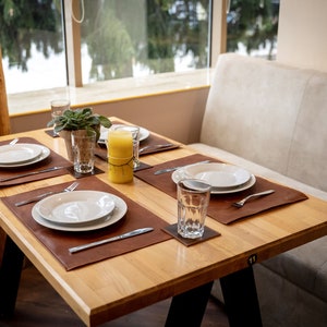 May include: A wooden table with four place settings, each with a white plate, brown leather placemat, silverware, and a glass. A yellow candle sits in the center of the table.