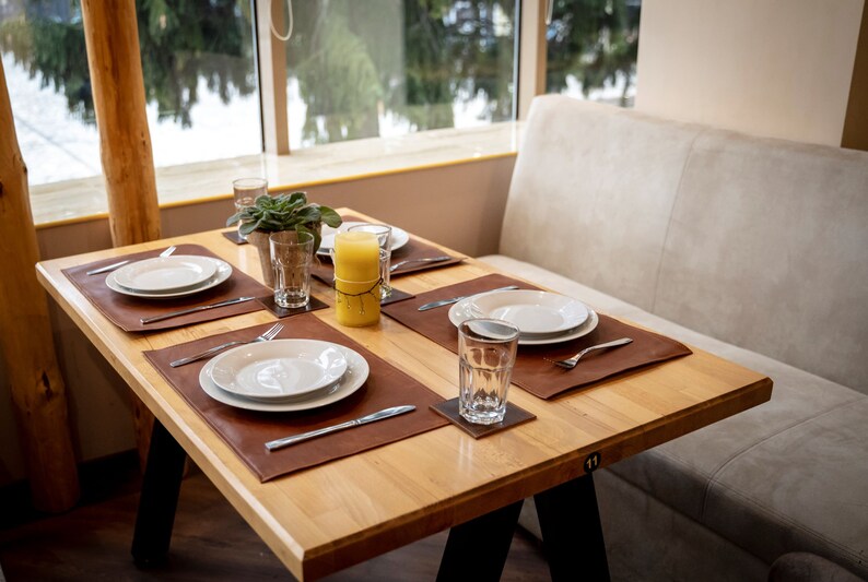 May include: A wooden table with four place settings, each with a white plate, brown leather placemat, silverware, and a glass. A yellow candle sits in the center of the table.
