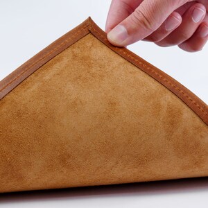 May include: A close-up of a brown leather coaster with a stitched edge. The coaster is folded in half, revealing the soft suede lining.