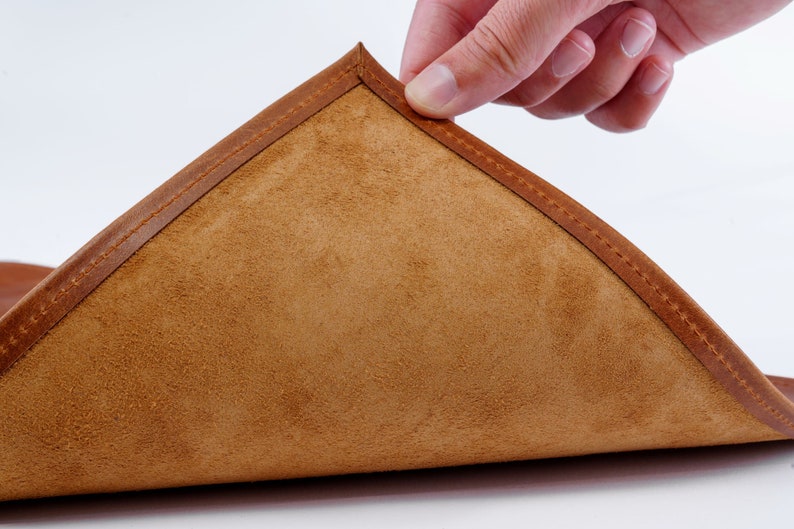 May include: A close-up of a brown leather coaster with a stitched edge. The coaster is folded in half, revealing the soft suede lining.
