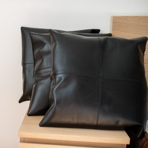 Pu&ograve; includere: Tre cuscini da divano in pelle nera con un design cucito. I cuscini sono disposti su una superficie in legno.