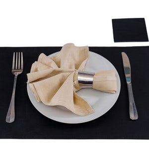 May include: A black placemat with a white plate, a beige napkin, a silver fork, and a silver knife. The napkin is folded and placed on the plate with a silver napkin ring.