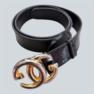 May include: A black leather belt featuring a large, circular, metallic buckle with a polished gold and silver finish. The buckle consists of two interlocking, stylized C-shaped components. The belt strap is smooth, dark, and coiled into a loop.