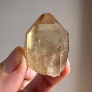 May include: A clear, light brown crystal with a pointed top and multiple facets. The crystal is translucent and has a slight yellow hue.