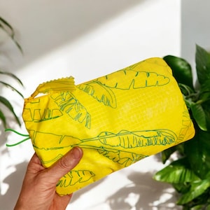 Può includere: Un involucro per alimenti riutilizzabile giallo con un motivo a foglie di banana verdi. L'involucro è piegato e tenuto in una mano.