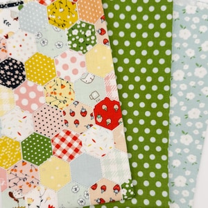 May include: A collection of fabric swatches in various patterns. One swatch features a patchwork design with hexagonal shapes in multiple colors. Other swatches include green polka dots and a light blue floral print. The fabrics are arranged on a white surface.