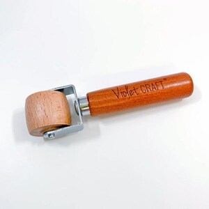 May include: A seam roller with a light-colored wooden cylindrical wheel, a silver metal frame, and a smooth, medium-brown wooden handle. The handle features the text Violet CRAFT branded into the wood surface in a dark, cursive font.
