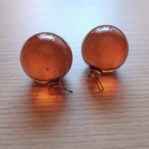 May include: Two amber-colored glass Christmas ornaments. Each ornament is a sphere with a small metal cap and wire loop for hanging. The ornaments are resting on a light-colored wooden surface.
