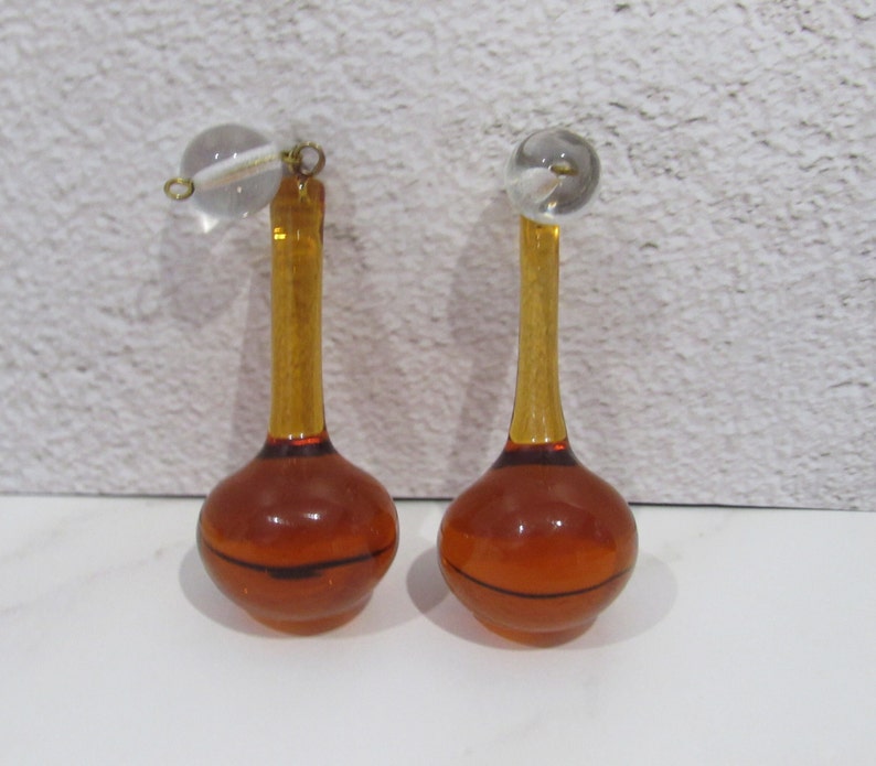 May include: A pair of amber glass bottle stoppers with clear glass ball tops. The stoppers have a thin, dark line around the bottom of the bulb.