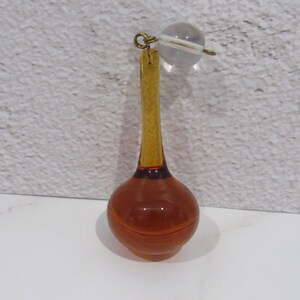 May include: A glass bottle stopper with a long, amber-colored neck and a round, amber-colored bulb. The stopper is topped with a clear glass ball.
