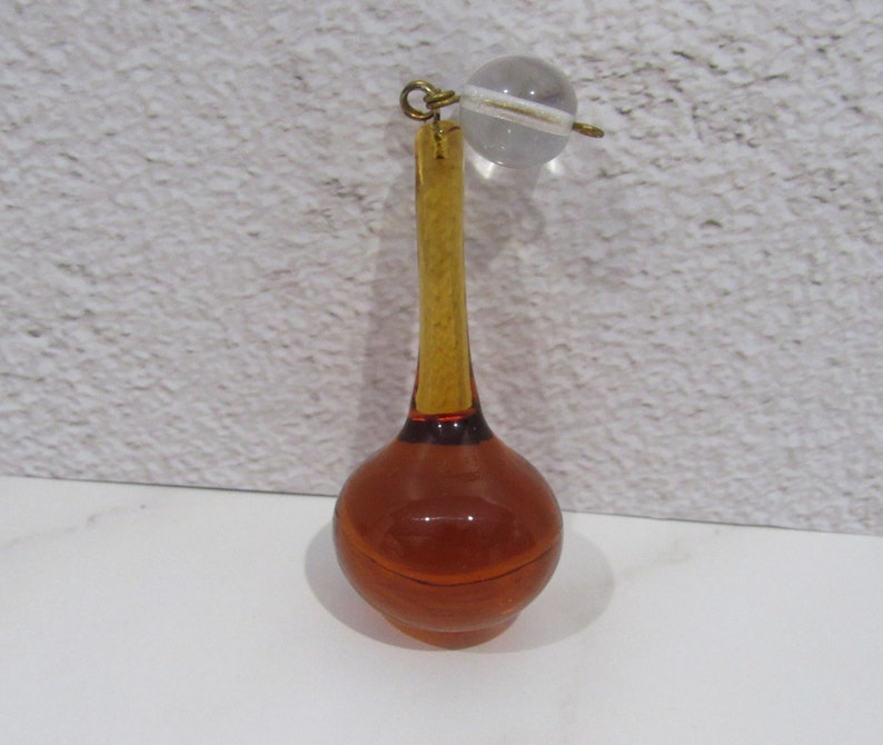 May include: A glass bottle stopper with a long, amber-colored neck and a round, amber-colored bulb. The stopper is topped with a clear glass ball.