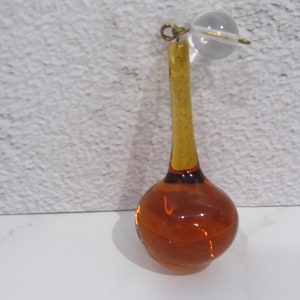 May include: A teardrop-shaped amber glass ornament with a clear glass ball at the top. The ornament has a gold-colored metal loop at the top.