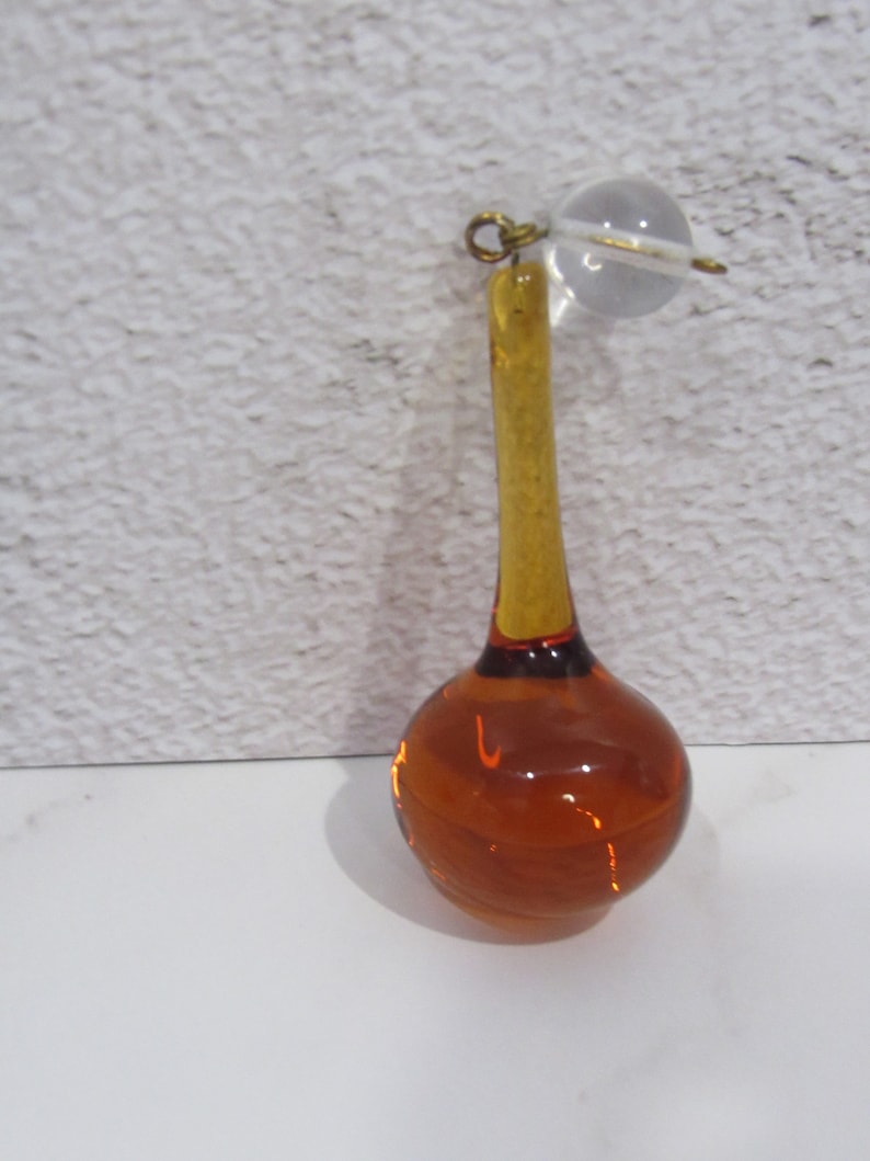 May include: A teardrop-shaped amber glass ornament with a clear glass ball at the top. The ornament has a gold-colored metal loop at the top.