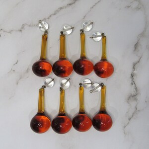 May include: Eight amber glass teardrop-shaped chandelier prisms with silver caps. The prisms are hanging from a white surface.