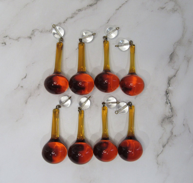 May include: Eight amber glass teardrop-shaped chandelier prisms with silver caps. The prisms are hanging from a white surface.
