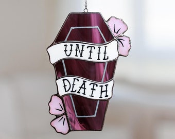 Puede incluir: Un adorno de ventana de vidrio tintado con forma de ataúd con una pancarta blanca que dice "Until Death" y flores rosas a cada lado.