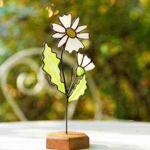 Puede incluir: Una escultura de flores de vidrio coloreado con piezas de vidrio blanco y verde. La flor está sobre una base de madera.