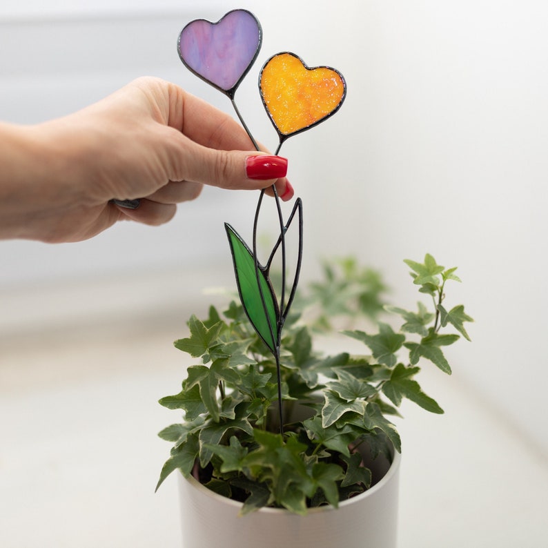 Op de afbeelding: Een hartvormige bloem van gekleurd glas met een paars en oranje hart op een zwarte draadsteel met een groen blad. De bloem staat in een witte pot met groene klimop.