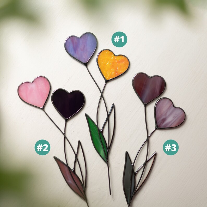 Op de afbeelding: Drie glas-in-lood hartvormige bloemdecoraties met verschillende gekleurde glazen harten. De harten zijn paars, roze, oranje en bruin. De bloemen zijn genummerd 1, 2 en 3.