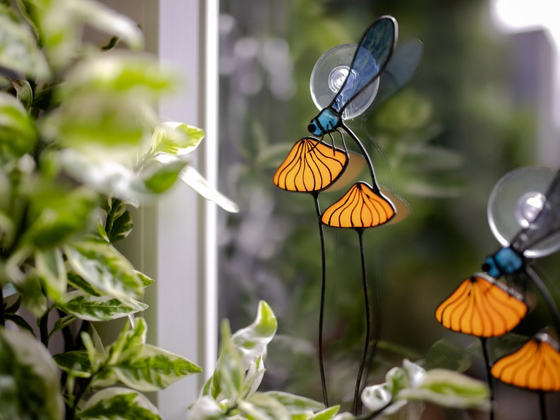 suncatcher in the form of two orange mushrooms with veins on which sits a dragonfly with blue wings in a plant vase. dragonfly stain glass