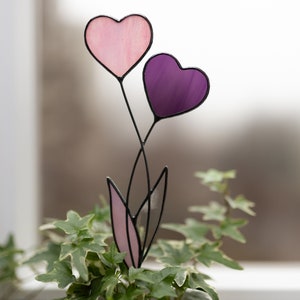 Op de afbeelding: Een hartvormige bloem van gekleurd glas met roze en paarse harten op zwarte stelen. De bloem staat in een pot met groene bladeren.