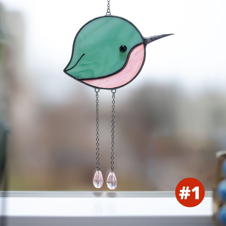 Suncatcher made with stained glass in the shape of a cartoon Hummingbird. Green hummingbird has a pink belly with long chain-legs