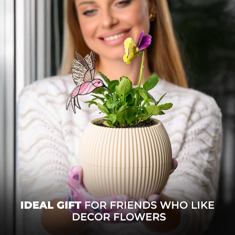 May include: A cream-colored, ribbed planter pot holding a flowering plant with a yellow and purple flower. A decorative pink hummingbird is attached to the plant. The text at the bottom reads: IDEAL GIFT FOR FRIENDS WHO LIKE DECOR FLOWERS.
