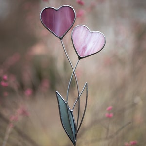 Op de afbeelding: Een glas-in-lood hartbloem met twee roze harten en een groene stengel. De harten zijn van glas en hebben een delicate, doorschijnende kwaliteit.