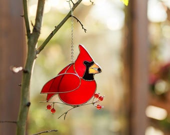 May include: A stained glass cardinal hanging from a branch with red berries. The cardinal is red with black eyes and a black beak. The berries are red and shiny.