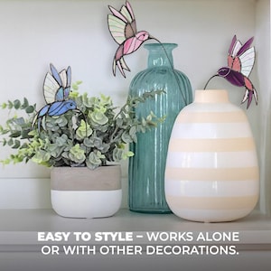 May include: Decorative hummingbird ornaments in shades of blue, pink, and purple, with delicate wire stems, are displayed near a green plant in a white and gray pot, and two decorative vases. The text reads: EASY TO STYLE - WORKS ALONE OR WITH OTHER DECORATIONS.
