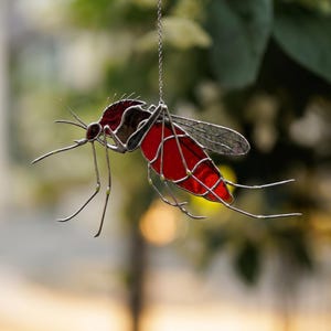 Op de afbeelding: Een gebrandschilderde glazen muggenvanger met een rood lichaam en transparante vleugels. Het insect hangt aan een zilveren ketting, met gedetailleerde draadpoten en antennes. De achtergrond is vervaagd met groen.