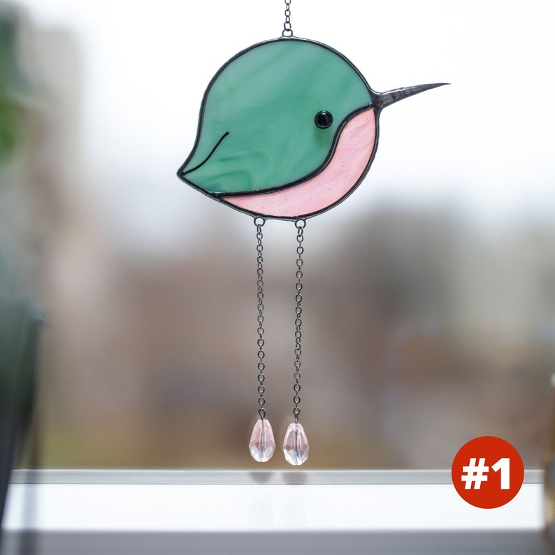 Suncatcher made with stained glass in the shape of a cartoon Hummingbird. Green hummingbird has a pink belly with long chain-legs