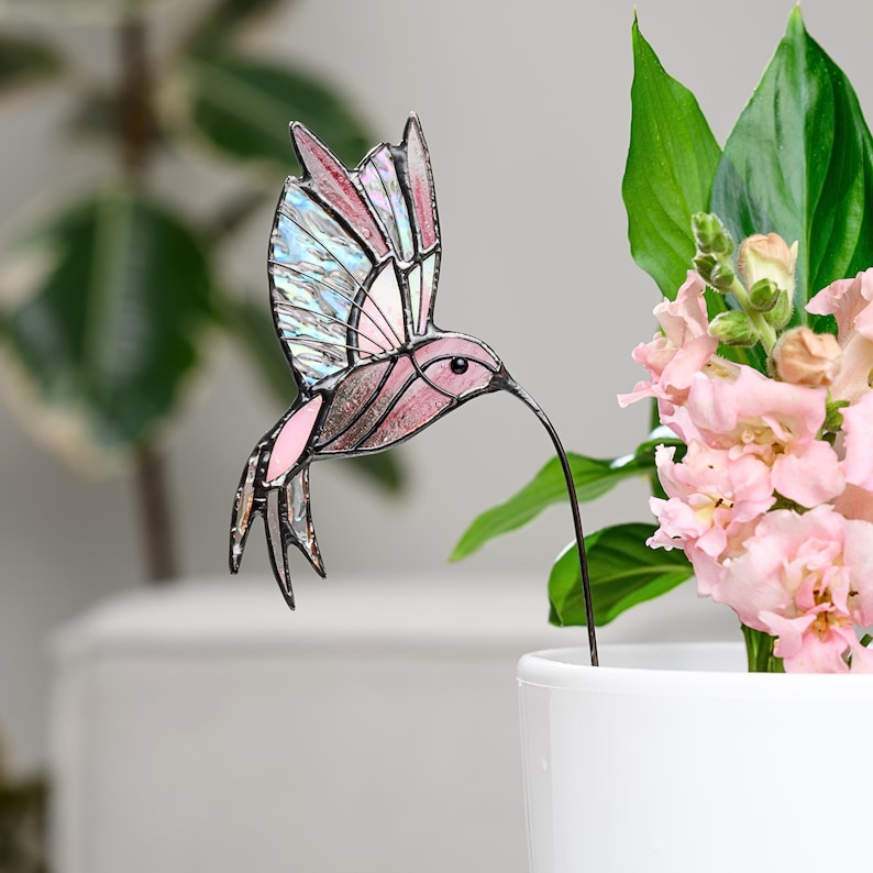 May include: A stained glass hummingbird decoration in shades of pink, brown, and blue. The bird has a long, thin beak and is attached to a black metal stake. The hummingbird is placed in a white pot with pink flowers and green leaves.