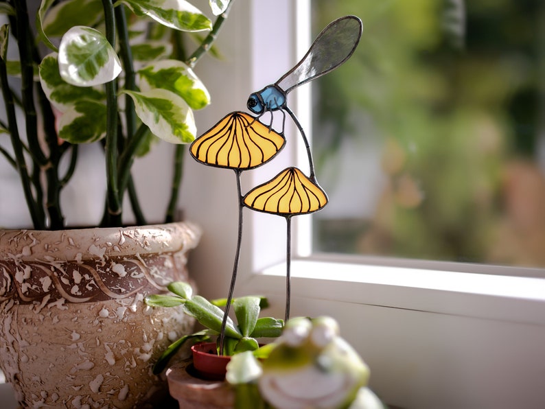 suncatcher in the form of two orange mushrooms with veins on which sits a dragonfly with blue wings in a plant vase