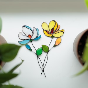 May include: A stained glass floral wall decor featuring two flowers, one blue and white and the other yellow and orange. The flowers are connected by black wire stems.