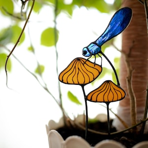 Peut inclure: Une libellule en verre coloré perchée sur deux décorations en forme de champignon jaune. La libellule est bleue avec des accents noirs. Les champignons sont jaunes avec des lignes noires.
