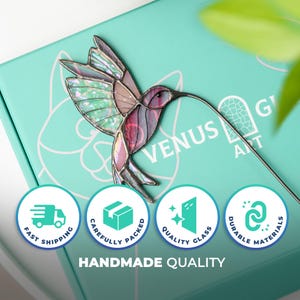 May include: A stained glass hummingbird with iridescent wings and a long beak. The bird is in shades of pink, purple, and green. The hummingbird is on a teal box with the words "Venus Art" and icons for fast shipping, quality glass, and durable materials.