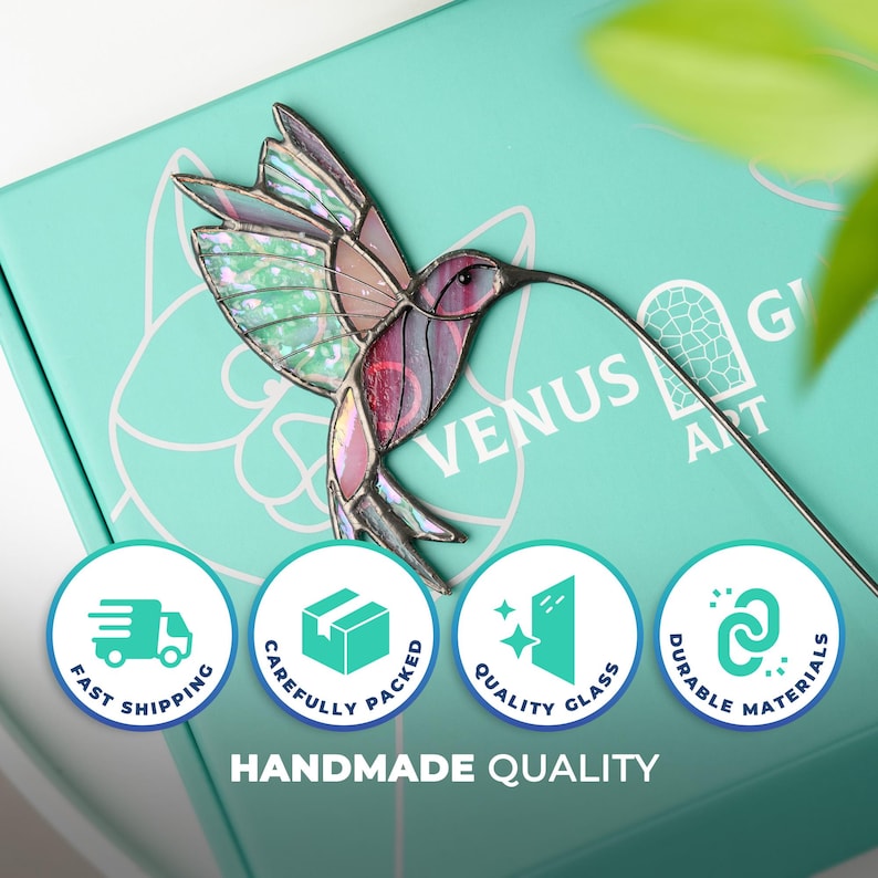 May include: A stained glass hummingbird with iridescent wings and a long beak. The bird is in shades of pink, purple, and green. The hummingbird is on a teal box with the words "Venus Art" and icons for fast shipping, quality glass, and durable materials.