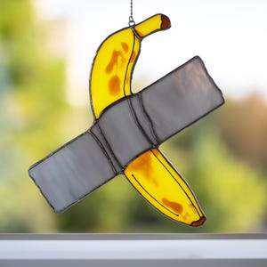Puede incluir: Una ventana de vitral colgante presenta una banana amarilla pegada a un trozo de cinta adhesiva gris.